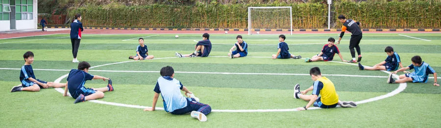 学生在人工草坪足球场上围成一圈做拉伸，教练站在一旁指导